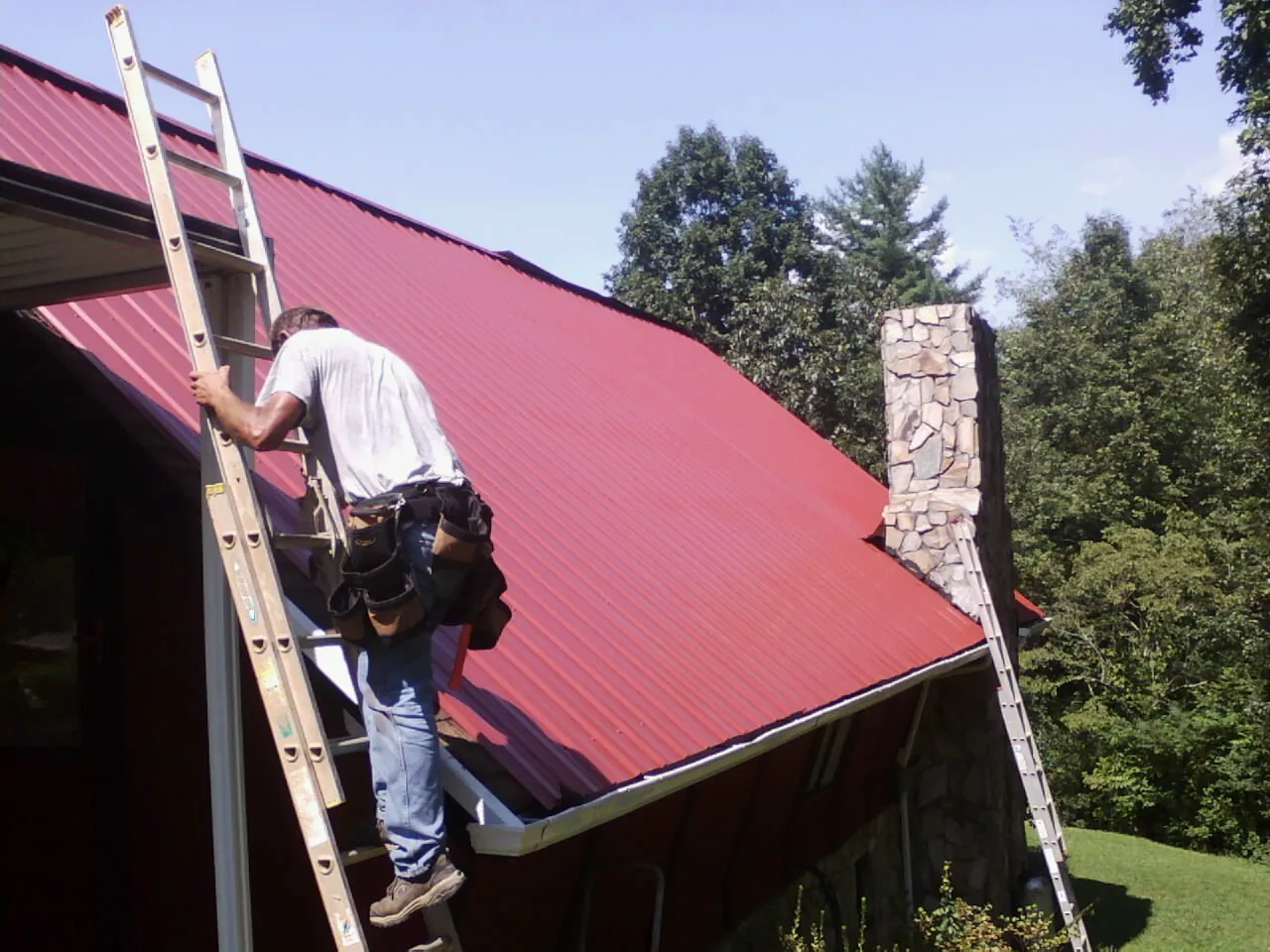 Roofer on a ladder beside a red metal roof and stone chimney — Soffit & Fascia Repair by Roofing Brothers in Newtown